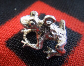 May include: A silver frog charm with a detailed design. The charm is on a red and black patterned fabric.