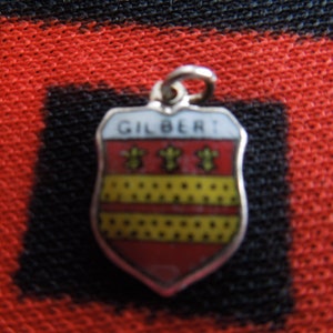 May include: Silver charm in the shape of a shield with the word "GILBERT" on it. The shield is red with yellow and black accents.