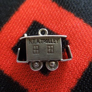 May include: Silver charm in the shape of a trolley car with the words "N.T.A. TROLLEY" on the side. The charm has two windows and two wheels.