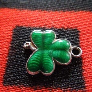 Puede incluir: Un amuleto de plata en forma de tr&eacute;bol verde con un dise&ntilde;o detallado. El amuleto tiene un anillo para sujetarlo a una pulsera o collar.