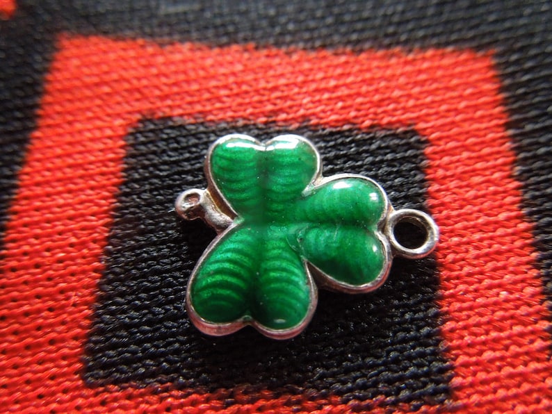 Puede incluir: Un amuleto de plata en forma de tr&eacute;bol verde con un dise&ntilde;o detallado. El amuleto tiene un anillo para sujetarlo a una pulsera o collar.