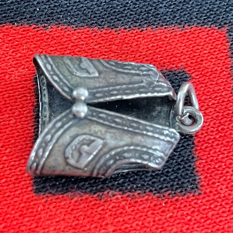 May include: A silver charm in the shape of a vest with intricate details and a loop for attaching to a bracelet or necklace.