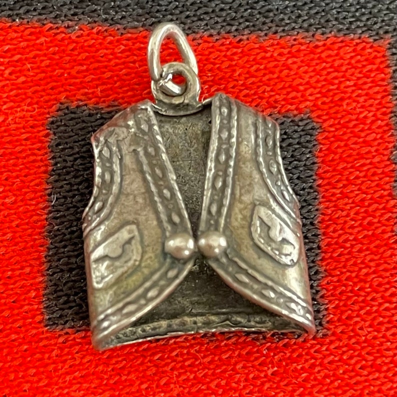 May include: A silver charm in the shape of a vest with a detailed design. The vest has a pointed collar and a button closure. The charm is made of metal and has a shiny finish.