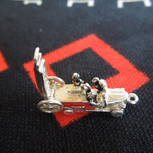 May include: Silver charm of a vintage car with two figures inside. The car has a red stripe and a raised hood. The charm has a loop for attaching to a bracelet or necklace.