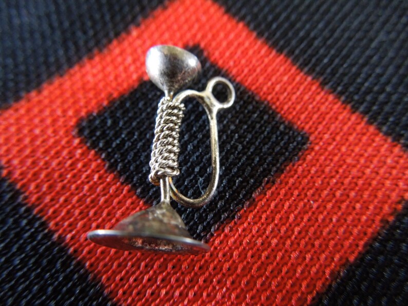 May include: A silver charm in the shape of a trumpet. The charm is detailed with a coiled design and a loop for attaching to a bracelet or necklace.