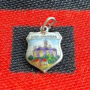 May include: A silver charm in the shape of a shield with a colourful image of a castle and the text "Schloss Schaumburg".