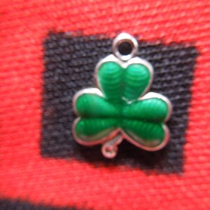 Puede incluir: Un amuleto de plata con forma de tr&eacute;bol verde. El amuleto tiene un bucle en la parte superior para sujetarlo a una pulsera o collar.