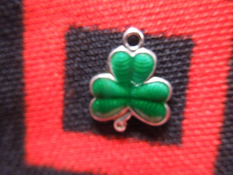 Puede incluir: Un amuleto de plata con forma de tr&eacute;bol verde. El amuleto tiene un bucle en la parte superior para sujetarlo a una pulsera o collar.