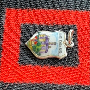 May include: A silver charm in the shape of a shield with a painted image of a castle and the text "Schloss Schaumburg".