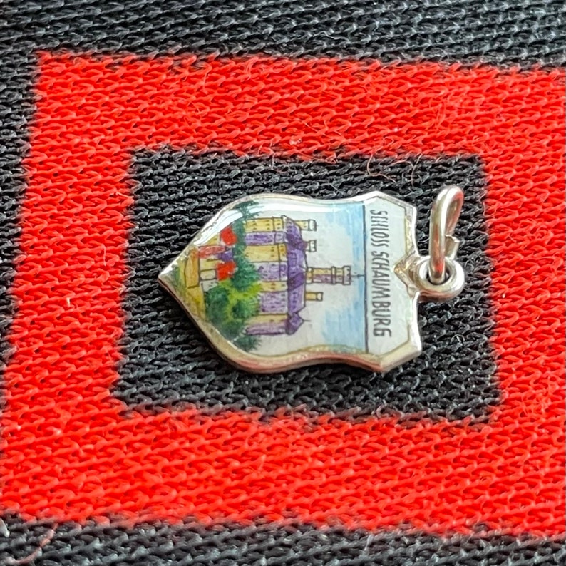 May include: A silver charm in the shape of a shield with a painted image of a castle and the text "Schloss Schaumburg".