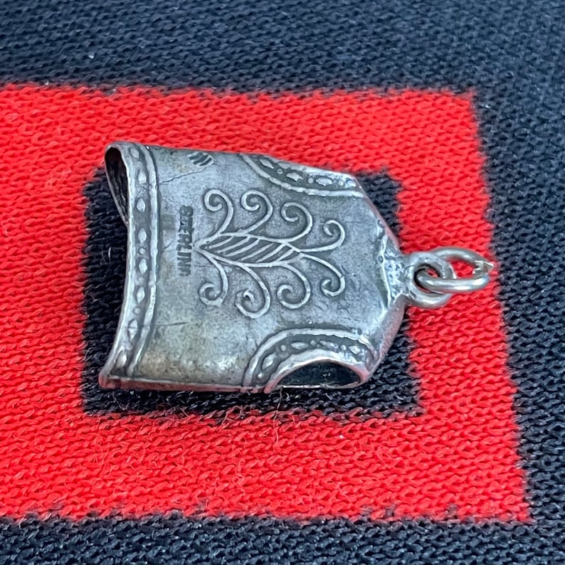May include: Silver charm in the shape of a suit of armor with a decorative design and the word "STERLING" inscribed on the back.