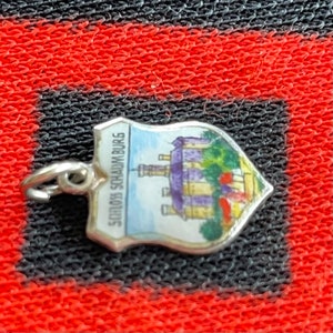 May include: A silver charm in the shape of a shield with a painted image of a castle and the text "Schloss Schaumburg".