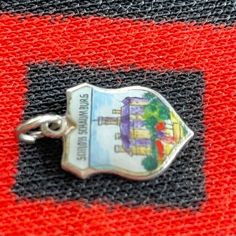 May include: A silver charm in the shape of a shield with a painted image of a castle and the text "Schloss Schaumburg".