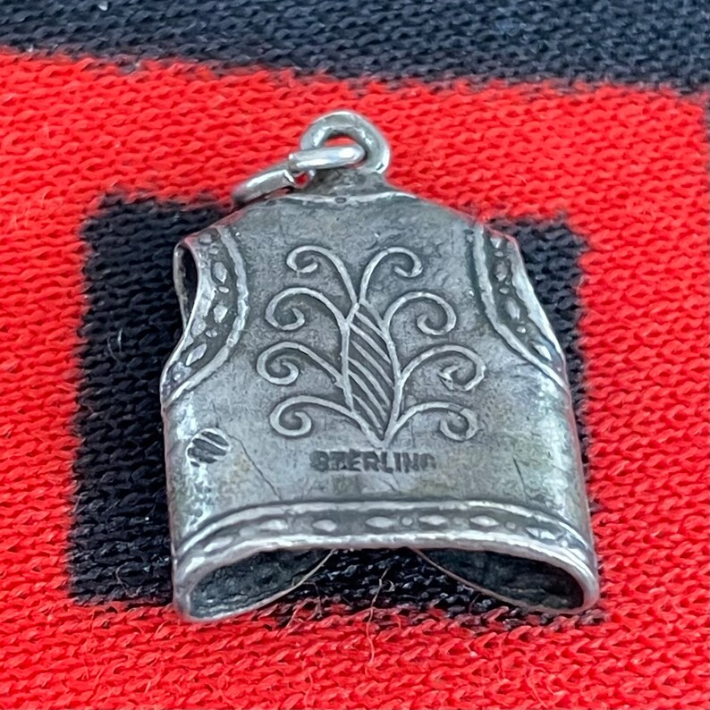 May include: Silver charm in the shape of a vest with a decorative design. The charm is marked "STERLING".