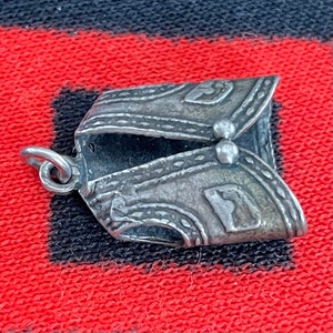 May include: A silver charm in the shape of a vest with intricate details and a small loop for attaching to a bracelet or necklace.