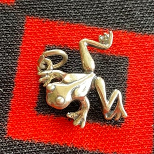 May include: A silver frog charm with a loop for attaching to a necklace or bracelet. The frog is in a sitting position with its legs outstretched.