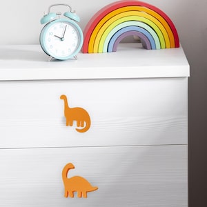 Könnte beinhalten: Ein hellblaues Wecker mit weißem Zifferblatt und silbernen Akzenten steht neben einem Holzspielzeug in Regenbogenform. Zwei orangefarbene Dinosaurier-Schubladengriffe sind an einer weißen Kommode befestigt. Das Regenbogenspielzeug hat Schichten aus Rot, Orange, Gelb, Grün, Blau und Lila.