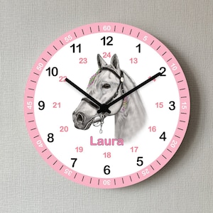 Könnte beinhalten: Eine weiße Uhr mit rosa Rand und schwarzen Zeigern. Das Zifferblatt der Uhr zeigt eine Zeichnung eines weißen Pferdes mit rosa Zaumzeug und einer rosa Blumenkrone. Der Name "Laura" ist unter dem Pferd gedruckt. Die Zahlen auf dem Zifferblatt sind in Schwarz und Rosa.