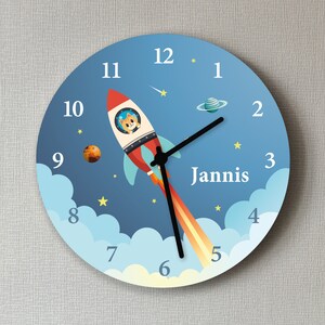 Könnte beinhalten: Eine blaue und weiße Kinderuhr mit einer Rakete und Planeten. Das Zifferblatt der Uhr hat die Zahlen 1 bis 12 und den Namen "Jannis" darauf geschrieben.