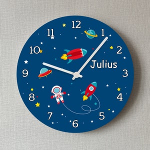 Könnte beinhalten: Eine blaue runde Uhr mit weißen Zeigern und schwarzen Zahlen. Die Uhr hat ein Weltraumthema mit einer Rakete, einem Astronauten und Sternen. Der Name "Julius" ist auf der Uhr geschrieben.
