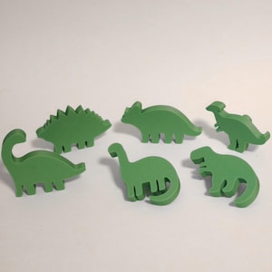 Könnte beinhalten: Sechs grüne Dinosaurier-Figuren in verschiedenen Formen sind auf weißem Hintergrund angeordnet. Die Sammlung umfasst einen Stegosaurus, einen Brontosaurus und einen Tyrannosaurus Rex. Diese Holzspielzeuge sind ideal für Kinder.