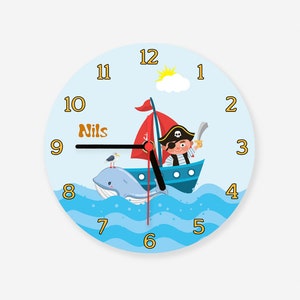 Könnte beinhalten: Eine blaue und weiße Kinderuhr mit einem Piraten auf einem Boot, einem Wal und einer Möwe. Das Zifferblatt der Uhr hat die Zahlen 1 bis 12 und den Namen "Nils" darauf geschrieben.