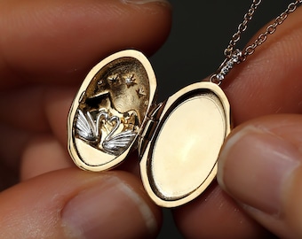 May include: A gold locket necklace with a delicate chain. The locket is open, revealing a small, detailed design of two swans inside.