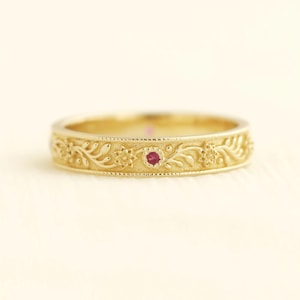 以下が含まれることがあります： 精巧なフローラルデザインのゴールドカラーのリング。リングの中央には、小さく丸い赤い宝石がセットされています。バンドには、花と葉の繊細でエレガントな彫刻が施されています。