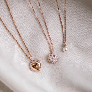 May include: Three gold necklaces with heart-shaped pendants. The pendant on the left has a small letter 'M' engraved inside. The pendant in the middle is a larger heart with a diamond-shaped stone in the center. The pendant on the right is a small pearl.