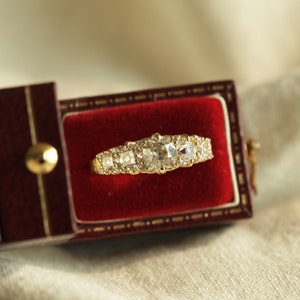 May include: A gold ring with a row of five round diamonds set in a band. The ring is in a red velvet ring box.