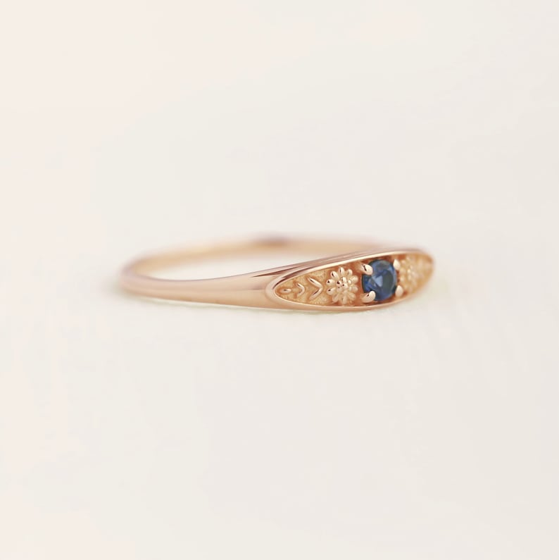 Pu&ograve; includere: Un delicato anello in oro rosa con una pietra preziosa blu circondata da dettagli floreali.