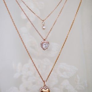 May include: Three gold necklaces with heart-shaped pendants. The top necklace has a small pearl pendant. The middle necklace has a heart-shaped pendant with a clear stone surrounded by smaller stones. The bottom necklace has a heart-shaped pendant with a smaller heart engraved inside.