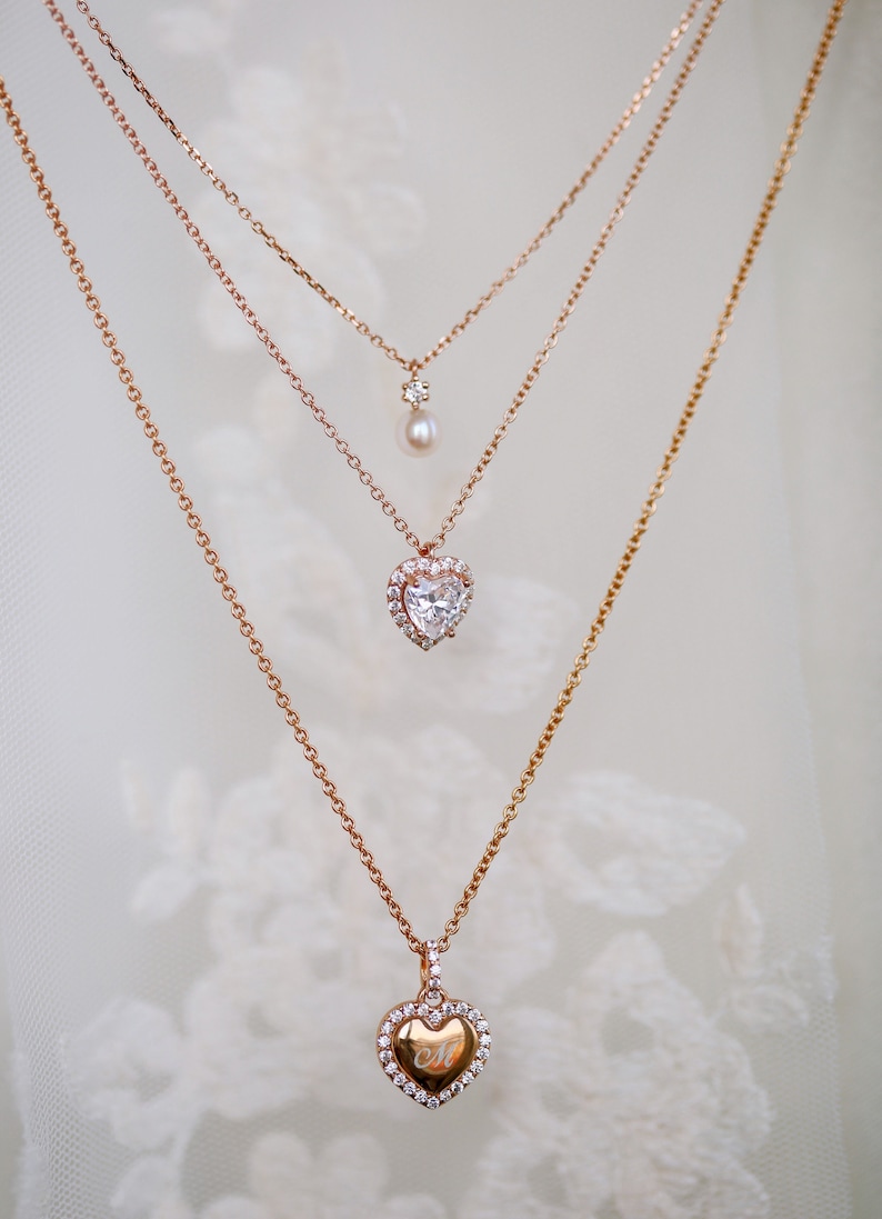 May include: Three gold necklaces with heart-shaped pendants. The top necklace has a small pearl pendant. The middle necklace has a heart-shaped pendant with a clear stone surrounded by smaller stones. The bottom necklace has a heart-shaped pendant with a smaller heart engraved inside.