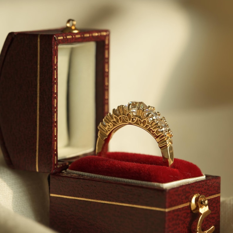 May include: A gold ring with a row of clear gemstones set in a decorative design, displayed in a red velvet lined box with a gold trim.