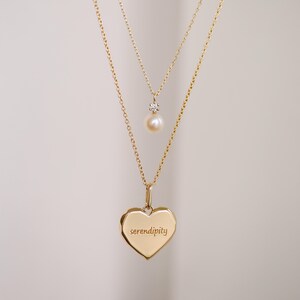 May include: A gold necklace with two pendants. The top pendant is a small pearl with a diamond accent. The bottom pendant is a heart-shaped charm with the word "serendipity" engraved on it.