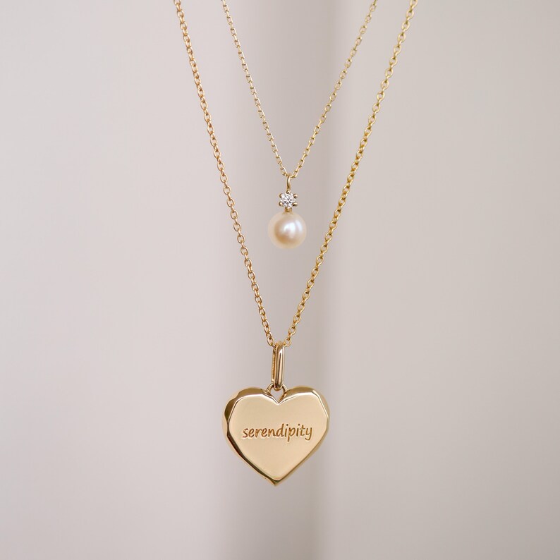 May include: A gold necklace with two pendants. The top pendant is a small pearl with a diamond accent. The bottom pendant is a heart-shaped charm with the word "serendipity" engraved on it.