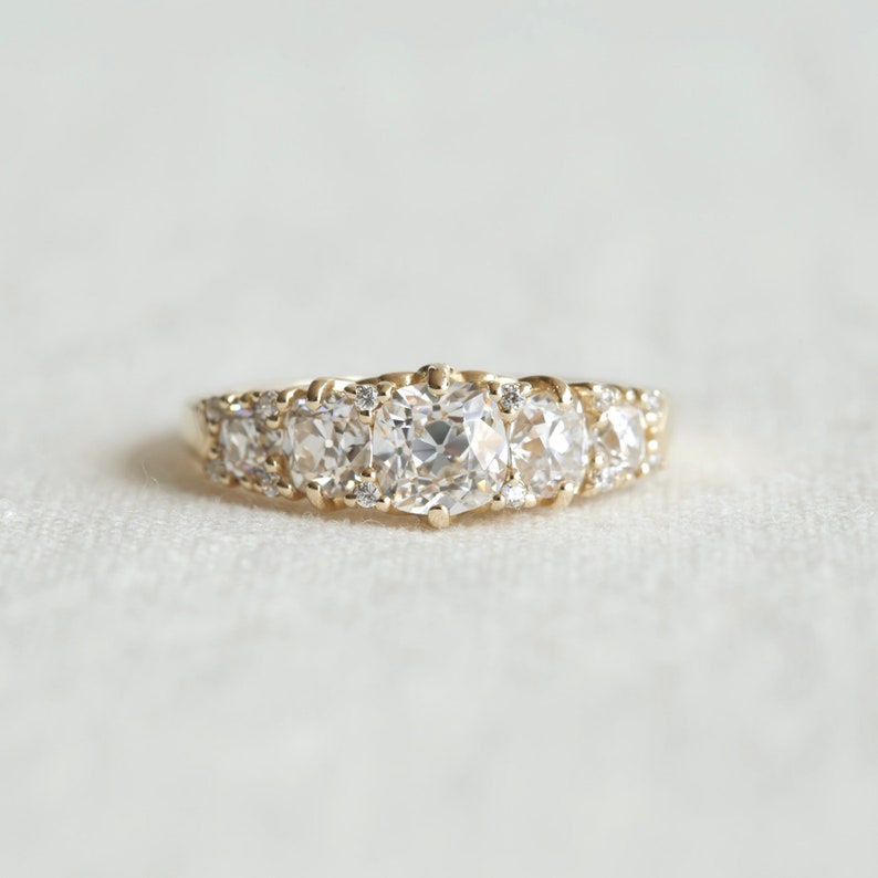 May include: A gold band ring with five round, clear gemstones. The center stone is larger than the other four stones.