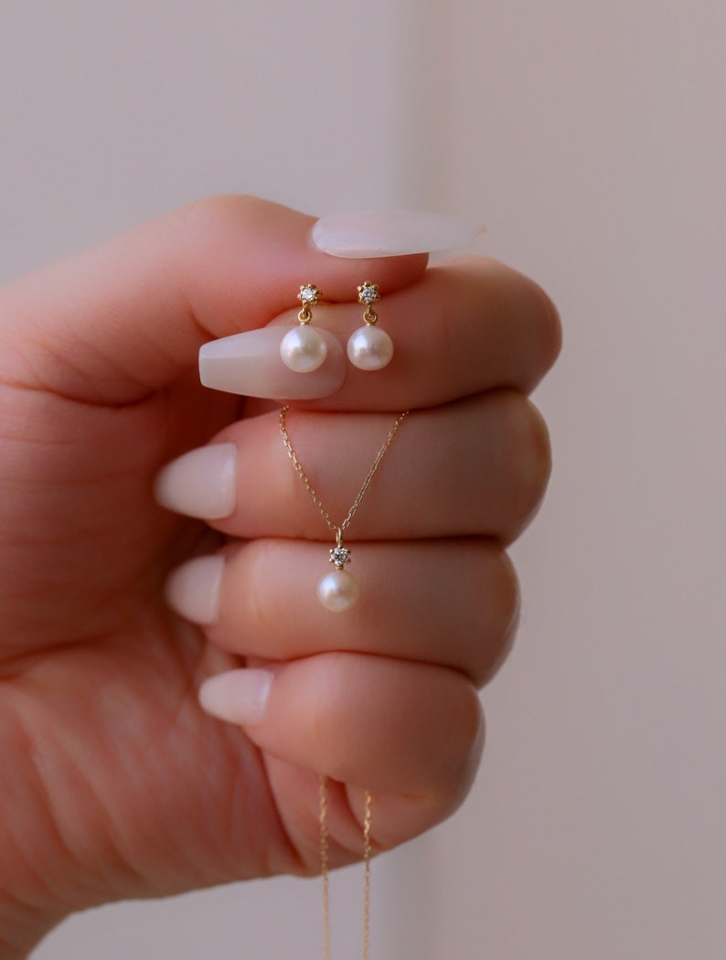 May include: A delicate gold necklace and earrings set featuring a small pearl and a tiny diamond accent. The necklace has a simple chain and the earrings are drop style.