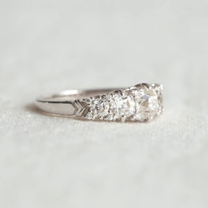 May include: A silver ring with a large, round, clear diamond in the center and smaller diamonds on either side. The ring is set on a white fabric background.