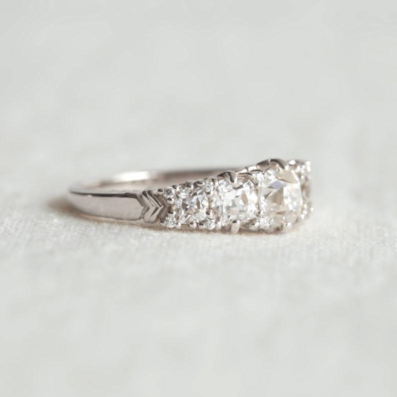 May include: A silver ring with a large, round, clear diamond in the center and smaller diamonds on either side. The ring is set on a white fabric background.