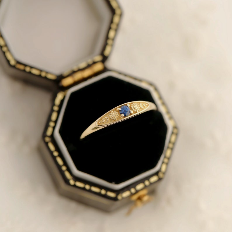 Pu&ograve; includere: Un anello d'oro con uno zaffiro blu e incisioni floreali, posto in una scatola per anelli nera.