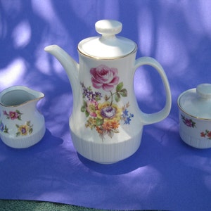 May include: A white porcelain tea set with a floral design. The set includes a teapot, creamer, and sugar bowl. The teapot has a gold trim and a floral design with pink, yellow, and purple flowers. The creamer and sugar bowl have a similar floral design.