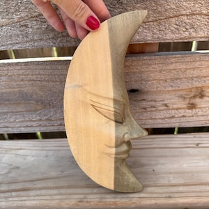 May include: A crescent moon wall carving made of light-colored wood. The piece features a carved face with closed eyes, a prominent nose, and a curved mouth. The wood grain is visible throughout the surface, showing natural variations in tone.