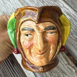 May include: A ceramic mug shaped like a jester's head with a glossy brown, yellow, and green finish. The face features detailed facial lines, dark eyebrows, and a subtle smile. The jester hat has pointed flaps with small red spheres at the ends.