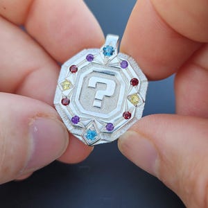 May include: A silver pendant with a question mark design at the center, surrounded by colorful gemstones. The pendant has a geometric shape with a loop at the top for a chain.