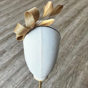 Op de afbeelding: Een gouden strik van stof met gouden bladaccenten op een witte mannequinkop. De strik is een haaraccessoire voor bruiloften of speciale gelegenheden.