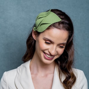 Puede incluir: Una mujer con un blazer blanco y un accesorio para el pelo de fieltro verde. El accesorio tiene forma de hoja y está colocado en el lado de su cabeza.