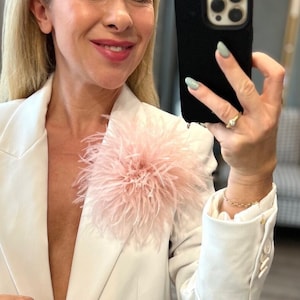 Op de afbeelding: Een wit blazer met een roze veren broche. De blazer heeft een enkele knoopsluiting en een revers.