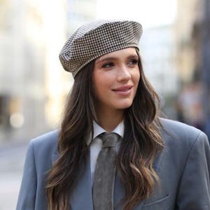 以下が含まれることがあります： グレーのブレザー、白いシャツ、柄物のベレー帽を着用した女性。ベレー帽は白と黒のチェック柄です。グレーのネクタイがコーディネートを完成させています。背景はぼやけています。