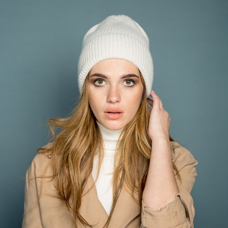 May include: A woman with long blonde hair wearing a white knit beanie and a beige trench coat.